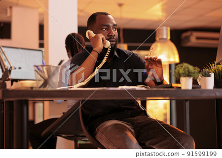 Company employee answering landline phone call to talk to manager about paperwork report on computer. Chatting on office telephone line, having remote conversation about business documents. Company employee answering landline phone call to talk to manager about paperwork report on computer. Chatting on office telephone line, having remote conversation about business documents. 91599199