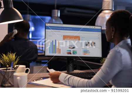 Female freelancer working with statistics charts on computer monitor, analyzing company data on paperwork. Using online information to create research report before deadline, at night after hours. 91599201