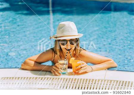 Portrait of beautiful cute smiling young woman lady girl in a bikini, hat, and sunglasses holding tropical lemonade cocktail while chilling in swimming pool. Hello summer holiday vacation. 91602840
