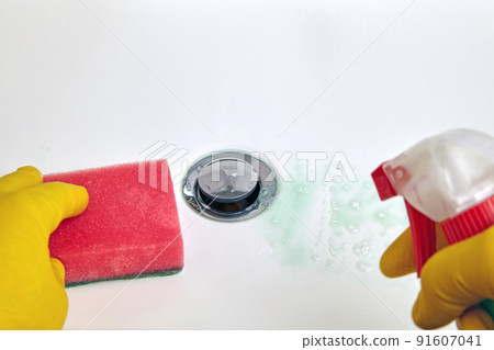 Person in yellow rubber gloves holds a sponge and a cleaning agent for cleaning the washbasin in the bathroom 91607041