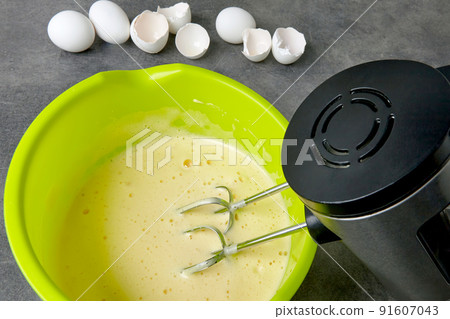 Beaten eggs with sugar in a green bowl with a beater immersed in a mixer and eggshells from chicken eggs Beaten eggs with sugar in a green bowl with a beater immersed in a mixer and eggshells from chicken eggs 91607043