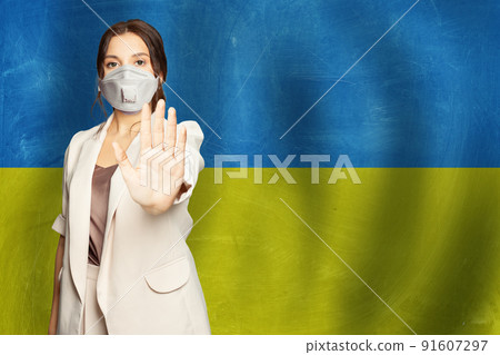 Portrait of serious young woman in protective mask against Ukrainian flag background 91607297
