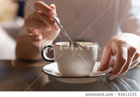 Closeup of female hands with french manicure holding cozy ceramic white mug of tea or coffee. Relax and comfort at home, cafe. Drinking hot cocoa. Empty space for text on blurry background, backplate 91611533