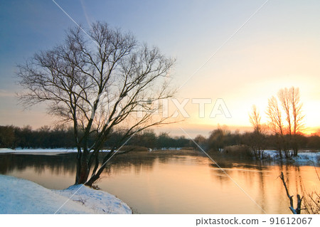 winter landscape with a lone tree with roots in ice, a road to a tree with ice waves on the snow against the backdrop of a bay with a blue sea in ice and a yellow-blue sunset in winter. winter landscape with a lone tree with roots in ice, a road to a tree with ice waves on the snow against the backdrop of a bay with a blue sea in ice and a yellow-blue sunset in winter. 91612067