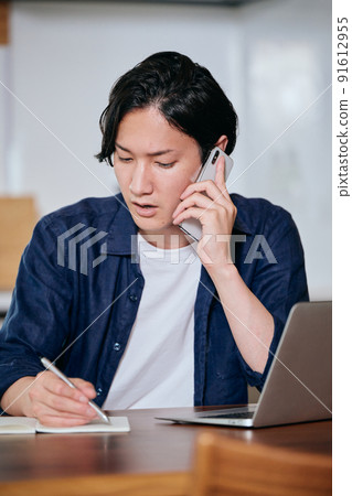 A young man taking notes in a notebook while making a phone call on his smartphone 91612955