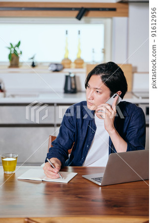 A young man taking notes in a notebook while making a phone call on his smartphone 91612956