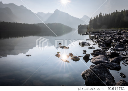 Still alpine lake reflecting sun and surrounding mountains and woods, Jasper NP, Canada 91614781