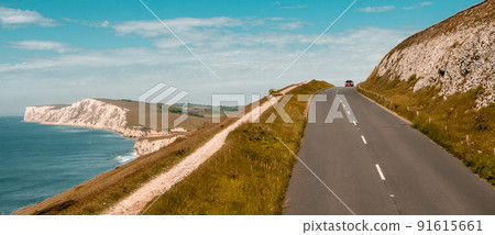 The Isle of WIght, UK. Road and white cliff of the Needles. Beautiful land view of white rocks and blue sea. Hills with car on road The Isle of WIght, UK. Road and white cliff of the Needles. Beautiful land view of white rocks and blue sea. Hills with car on road 91615661