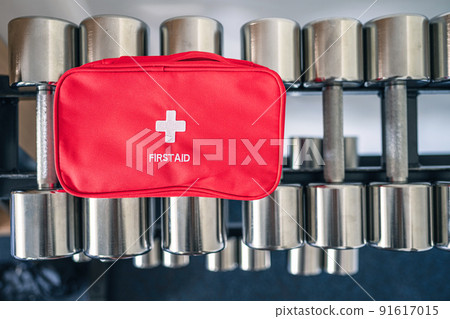 First aid kit red box on iron dumbbells in the fitness gym opposite the sport equipment and  jogging simulators. Healthy lifestyle, safety and help concept 91617015