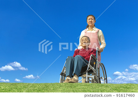 A middle-aged woman who takes care of an elderly woman in a wheelchair against the background of the blue sky. Old age, second life, old age, long-term care image 91617460