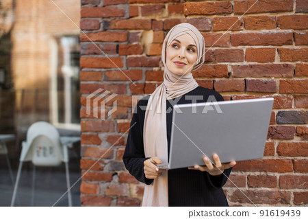 Portrait of smiling muslim woman relaxing using digital laptop while standing near red brick wall. 91619439