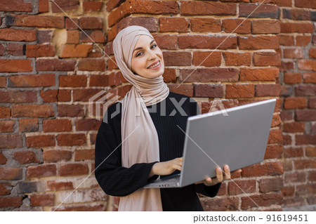 Portrait of smiling muslim woman relaxing using digital laptop while standing near red brick wall. Portrait of smiling muslim woman relaxing using digital laptop while standing near red brick wall. 91619451