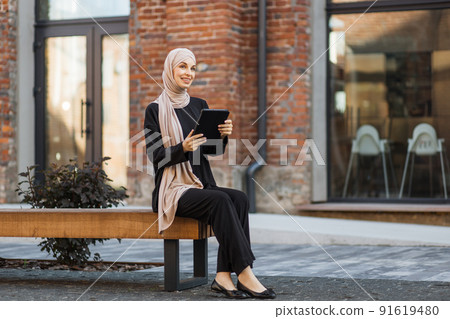 Pretty smiling muslim business woman in hijab sitting on bench and using tablet during video chat. 91619480