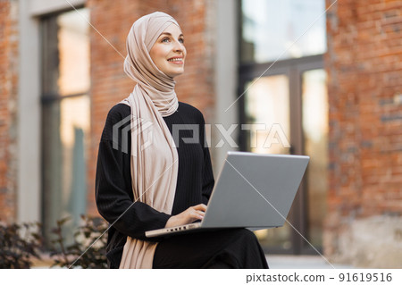 Pretty smiling muslim business woman in hijab sitting on bench and using laptop during video chat. Pretty smiling muslim business woman in hijab sitting on bench and using laptop during video chat. 91619516