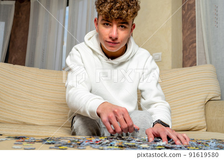 Caucasian young man with curly hair sitting on sofa at home assembling 1000 piece puzzle. Caucasian young man with curly hair sitting on sofa at home assembling 1000 piece puzzle. 91619580