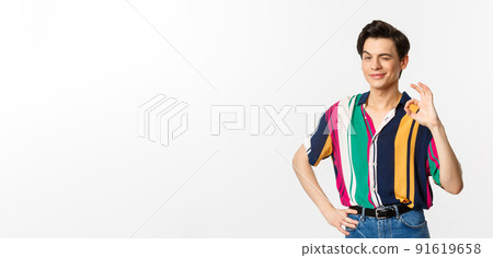 Confident young androgynous man showing okay sign, approve and agree, smiling and winking at camera, guarantee and recommend, standing over white background 91619658
