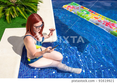 young redhead woman in bikini, looking at her smart phone sitting inside the pool. young girl on holiday sharing the moments on her social media. concept summer and free time. young redhead woman in bikini, looking at her smart phone sitting inside the pool. young girl on holiday sharing the moments on her social media. concept summer and free time. 91619921