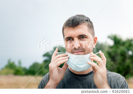 Portrait of a man wearing a medical blue mask due to an outbreak of a pandemic infection, disease or quarantine in a city park. Lifestyle during social distancing Coronavirus concept 91620081