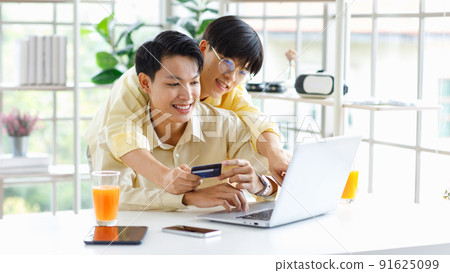Two Asian young happy pride teenager male gay men lover couple partner sitting smiling hugging cuddling together in front laptop notebook computer browsing surfing using credit card shopping online 91625099