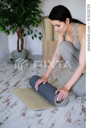 Young beautiful woman rolls up a yoga mat. Bright studio for meditation and yoga. Young beautiful woman rolls up a yoga mat. Bright studio for meditation and yoga. 91628226