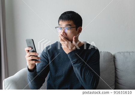 Portrait of excited asian man looking at mobile phone, man receiving good news using smartphone at home. Portrait of excited asian man looking at mobile phone, man receiving good news using smartphone at home. 91631610