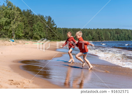 cute caucasian boys wearing red hoodies and blue underpants running from waves in lake with enthusiasm. cute caucasian boys wearing red hoodies and blue underpants running from waves in lake with enthusiasm. 91632571