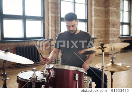 Young bearded man sitting at drum kit and performing on drums at studio during rehearsal 91633453