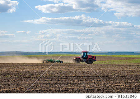 A red farm tractor in a cloud of dust cultivates the soil in the field with a cultivator after harvest. Summer sunny day. Fertile land. Modern agricultural machinery. Copy space. A red farm tractor in a cloud of dust cultivates the soil in the field with a cultivator after harvest. Summer sunny day. Fertile land. Modern agricultural machinery. Copy space. 91633840