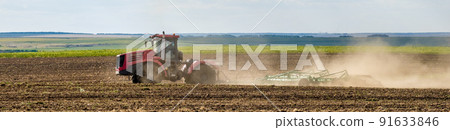 A red farm tractor in a cloud of dust cultivates the soil in the field with a cultivator after harvest. Summer sunny day. Fertile land. Modern agricultural machinery. Copy space. A red farm tractor in a cloud of dust cultivates the soil in the field with a cultivator after harvest. Summer sunny day. Fertile land. Modern agricultural machinery. Copy space. 91633846