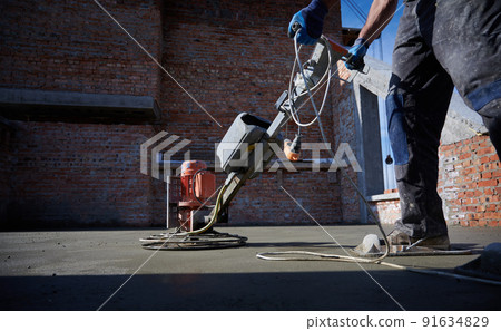 Close up male builder hands in protective gloves holding grout-grinding machine on concrete surface on the background of brick wall outside. 91634829
