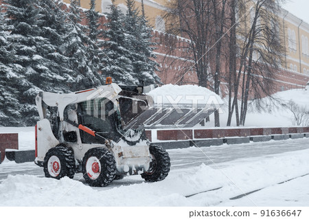 A small loader excavator bobcat removes snow from the sidewalk near the Kremlin walls during a heavy snowfall. A small loader excavator bobcat removes snow from the sidewalk near the Kremlin walls during a heavy snowfall. 91636647