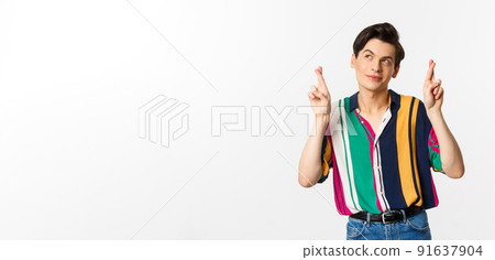 Dreamy young man making wish with fingers crossed, smiling and looking at upper left corner thoughtful, standing over white background 91637904