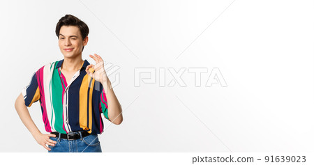 Confident young androgynous man showing okay sign, approve and agree, smiling and winking at camera, guarantee and recommend, standing over white background 91639023
