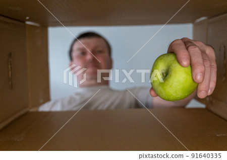 A man takes food out of a cardboard box. A green apple is taken out of the donation box. Bottom view. Inside view. Close-up. Selective focus. 91640335