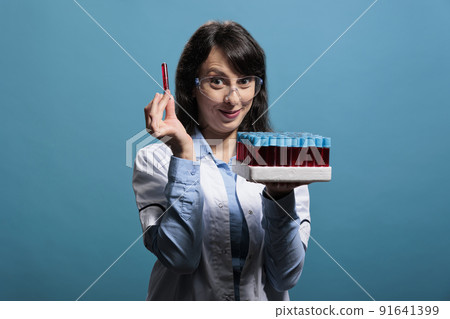 Cheerful biochemist expert analyzing laboratory glassware tray having test tubes filled with red liquid substance. Microbiology specialist having lab glass tubes filled with sample on blue background. Cheerful biochemist expert analyzing laboratory glassware tray having test tubes filled with red liquid substance. Microbiology specialist having lab glass tubes filled with sample on blue background. 91641399