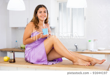 Portrait of young woman sitting on table in kitchen and drinking coffee 91644197