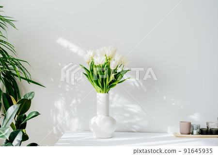 Scandinavian home interior with spring bouquet of white tulip flowers in ceramic vase standing on cabinet. Minimalist design with green plants and white wall with shadows. Biophilia style. Springtime. 91645935