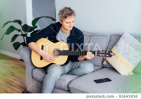 Man taking online guitar lessons via a mobile application on his phone at home sitting on the gray couch, sofa in the living room. He watching acoustic guitar tutorial. New hobby. Selective focus. 91645956