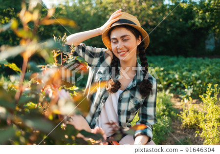 Portrait of smiling young woman in a straw hat sits in the garden, looking at flowers of a rose Portrait of smiling young woman in a straw hat sits in the garden, looking at flowers of a rose 91646304
