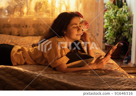 Smiling young woman is lying on bed and listening to music, putting headphones to her ear, looking into her smartphone. Sunset lighting from the window Smiling young woman is lying on bed and listening to music, putting headphones to her ear, looking into her smartphone. Sunset lighting from the window 91646316