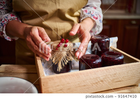 Woman hands next to a jar with jam, ornate with red currant berries on the burlap on the cover Woman hands next to a jar with jam, ornate with red currant berries on the burlap on the cover 91647410
