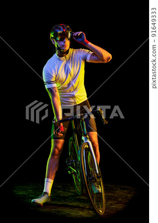 Full-length portrait of young male cyclist on bicycle in cycling shorts and protective helmet isolated on dark background in neon. Concept of sport, speed, energy 91649333