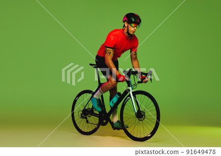Young sportsman, cyclist on bicycle in sports uniform and protective helmet training isolated on green background. Concept of active life, rest, travel, energy, sport Young sportsman, cyclist on bicycle in sports uniform and protective helmet training isolated on green background. Concept of active life, rest, travel, energy, sport 91649478