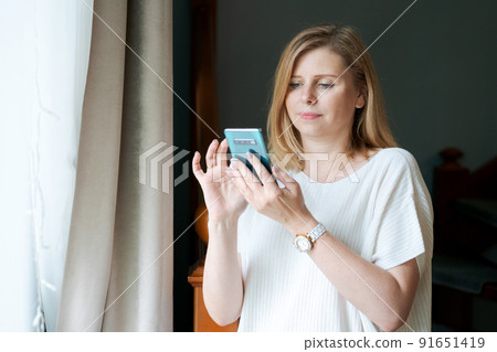 Happy adult woman talking on phone while standing at window against background Happy adult woman talking on phone while standing at window against background 91651419