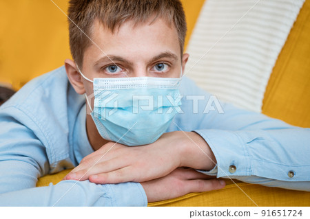 a young guy in a shirt and a protective mask sits on the couch a young guy in a shirt and a protective mask sits on the couch 91651724