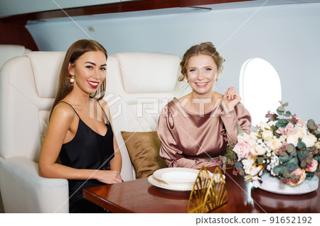 Two gorgeous caucasian women in dresses are sitting at a table in a private jet 91652192