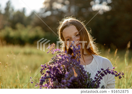 Close up portrait of young beautiful redhead woman with freckles, wearing white dress, posing in the nature. Girl with red hair holding flowers. Natural beauty. Diversity, individual uniqueness. 91652875
