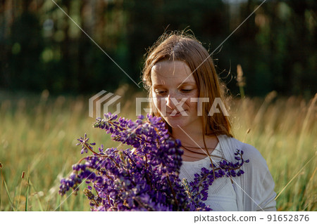Close up portrait of young beautiful redhead woman with freckles, wearing white dress, posing in the nature. Girl with red hair holding flowers. Natural beauty. Diversity, individual uniqueness. 91652876