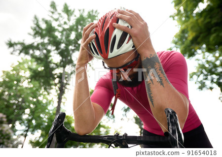 Tired male cyclist in sportswear, glasses and protective helmet walking with his bike in the forest to take a break after riding. Sky blue and forest in the background. Tired male cyclist in sportswear, glasses and protective helmet walking with his bike in the forest to take a break after riding. Sky blue and forest in the background. 91654018