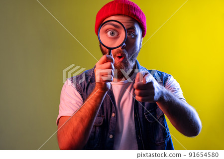 Bearded hipster man standing, holding magnifying glass and looking at camera with big zoom eye, pointing to camera with surprised face. Indoor studio shot isolated on colorful neon light background. Bearded hipster man standing, holding magnifying glass and looking at camera with big zoom eye, pointing to camera with surprised face. Indoor studio shot isolated on colorful neon light background. 91654580